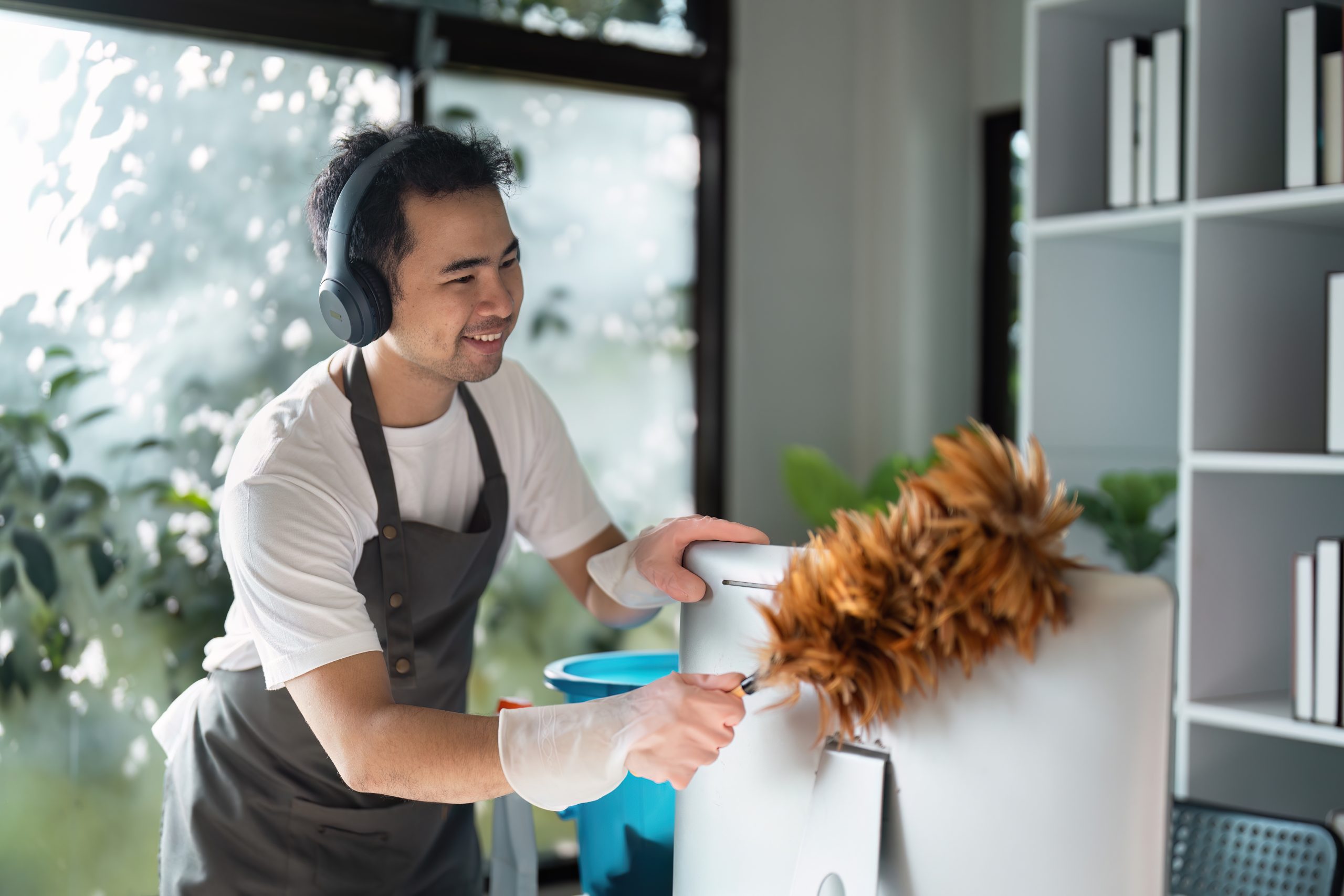 Smiling Man Cleaning Office Desk with Feather Duster and Headphones in Modern Workspace